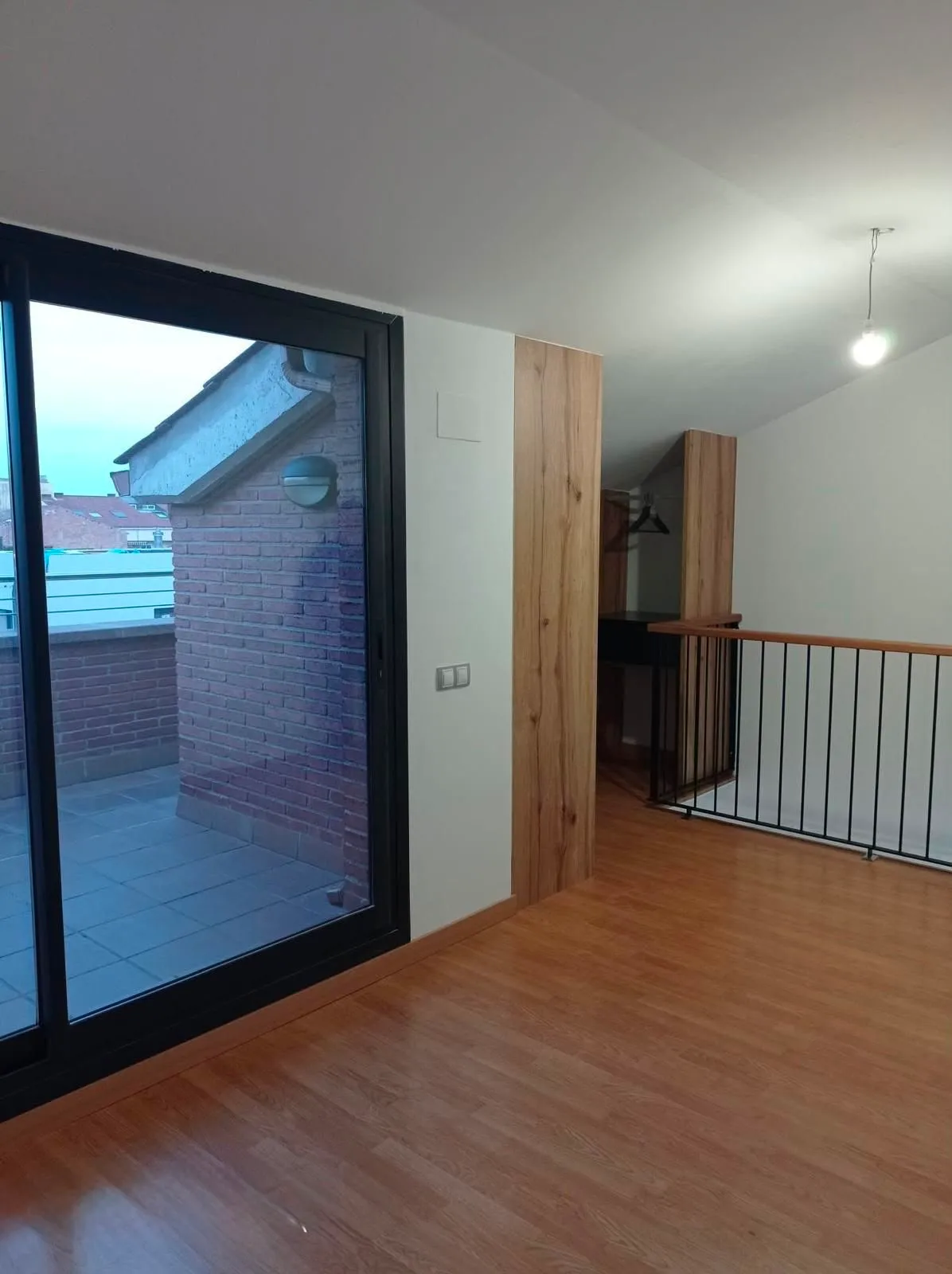 Upper-level room with wood floor, sliding glass door leading to a small brick terrace, wooden wall panels, metal railing overlooking a lower level, and a hanging ceiling light.