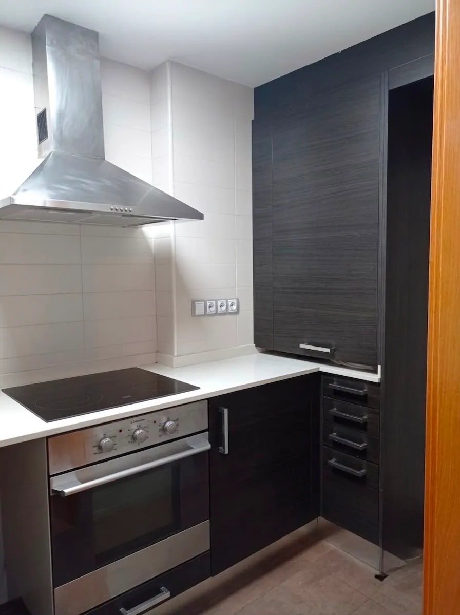 Kitchen corner with an electric cooktop, stainless steel oven, range hood, white countertop and dark wood cabinets.