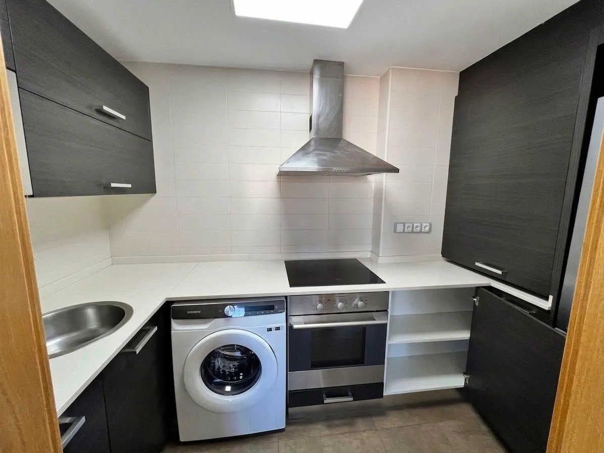 Kitchen with white countertop, stainless steel range hood, electric cooktop, built-in oven, front-loading washing machine, round sink, and dark wood cabinets.