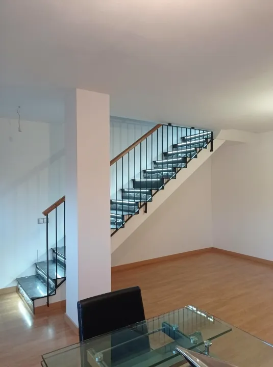 Open interior space with a staircase along a wall, glass table and chair in foreground, and wood-tone flooring.