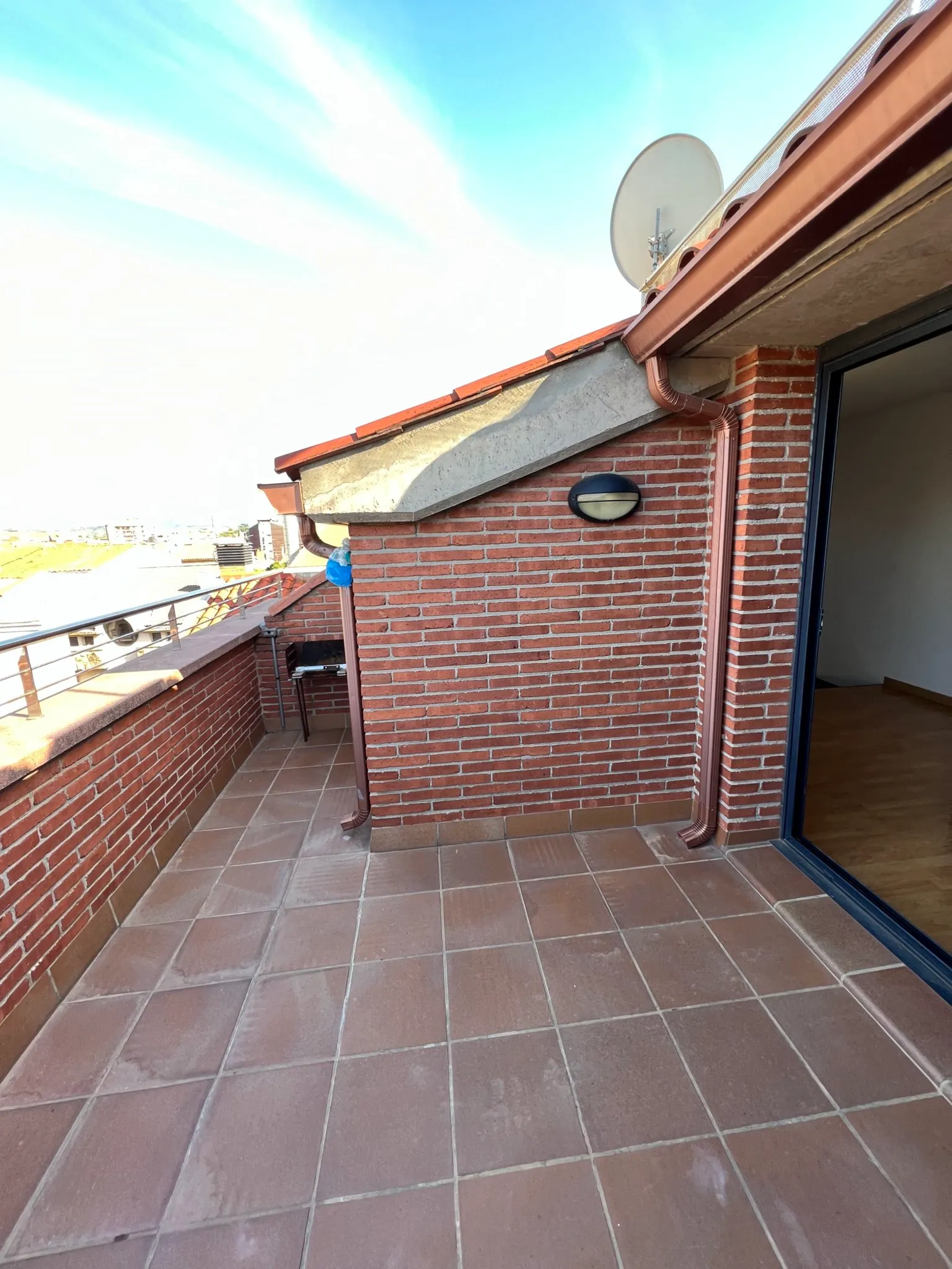 Balcony with tiled floor, low brick wall and metal railing, sloped roof with drainpipes, satellite dish and a wall light next to a sliding door