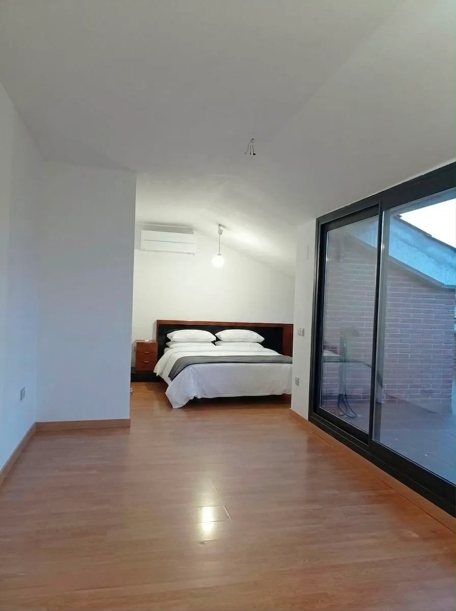 Bedroom with a double bed, two pillows, bedside table, wall-mounted air conditioning unit and large sliding glass doors to a balcony