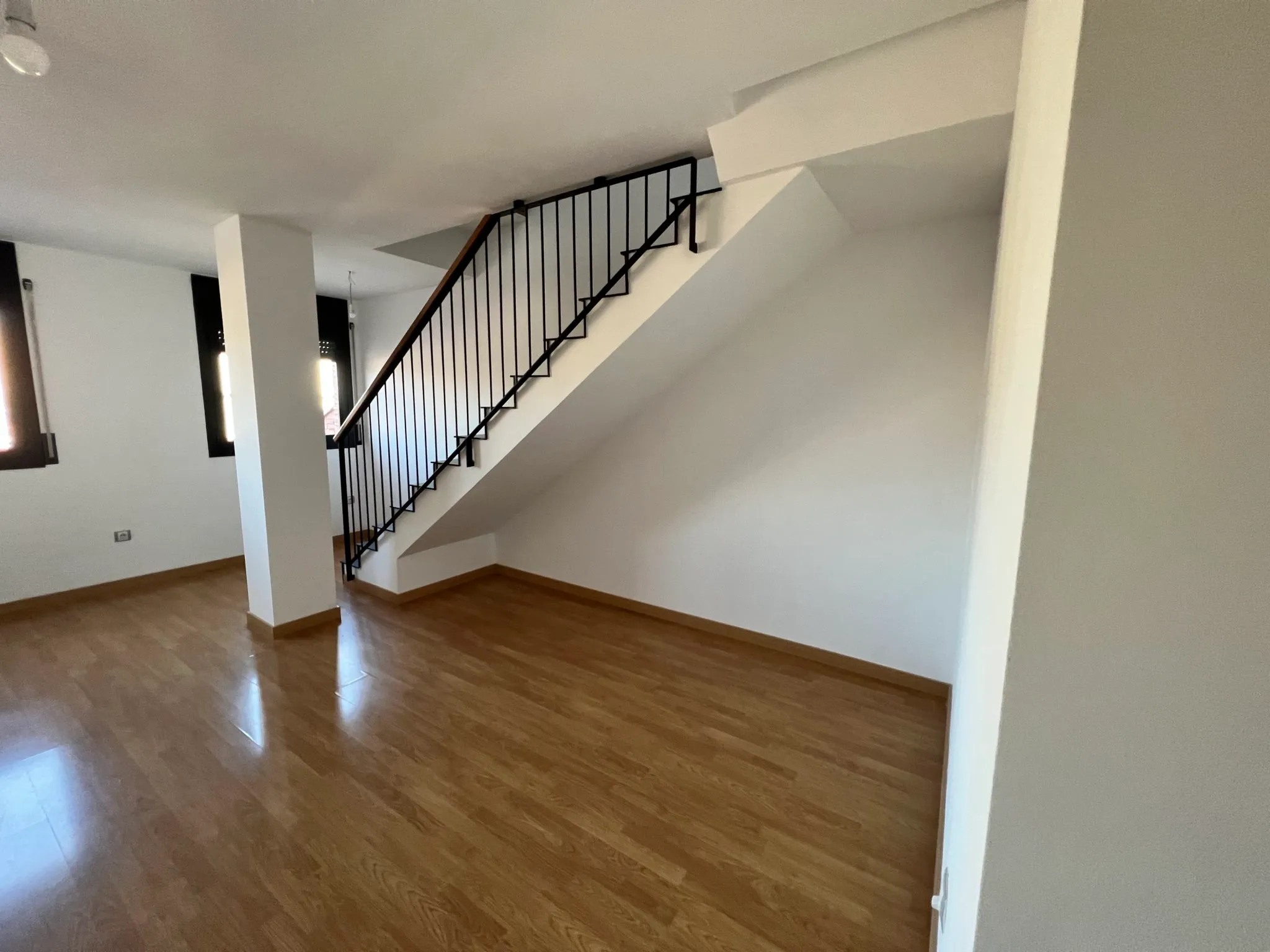 Open living area with a staircase and black metal railing, white walls, a support column, and wood-look flooring