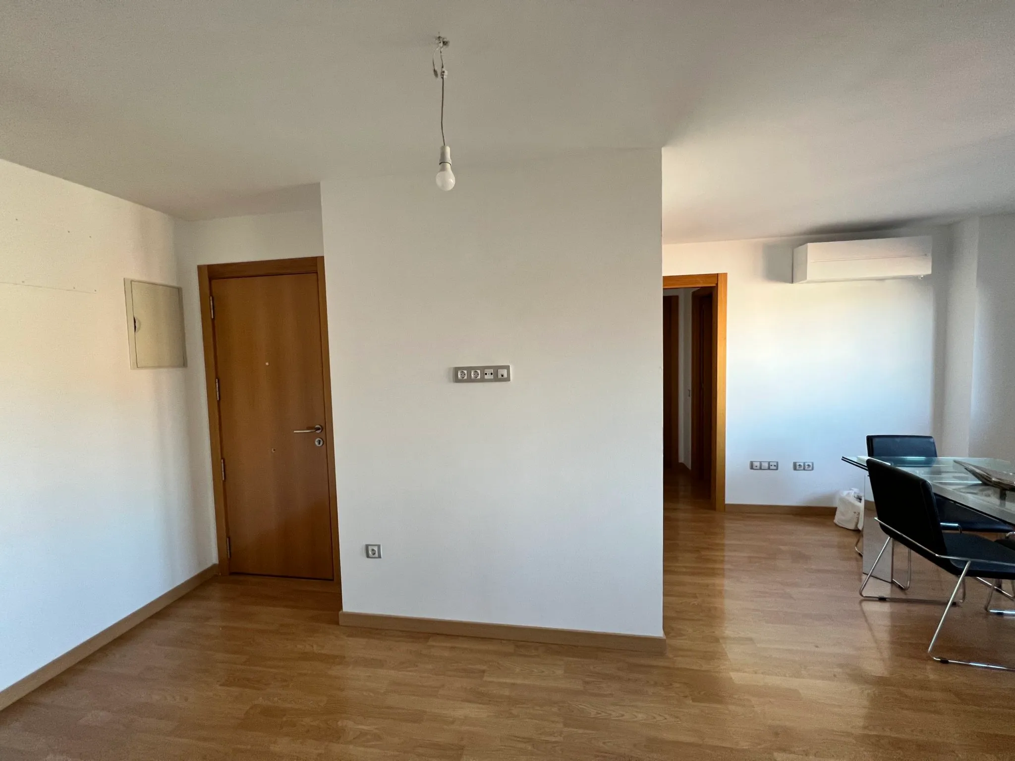 Open-plan living area with wood floor, white walls, a wooden door, visible air conditioning unit and a glass dining table with chairs at right
