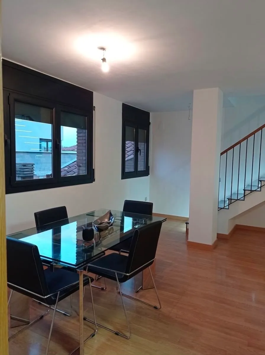 Dining room with a glass dining table, four black chairs, three windows on one wall and a staircase visible to the right.