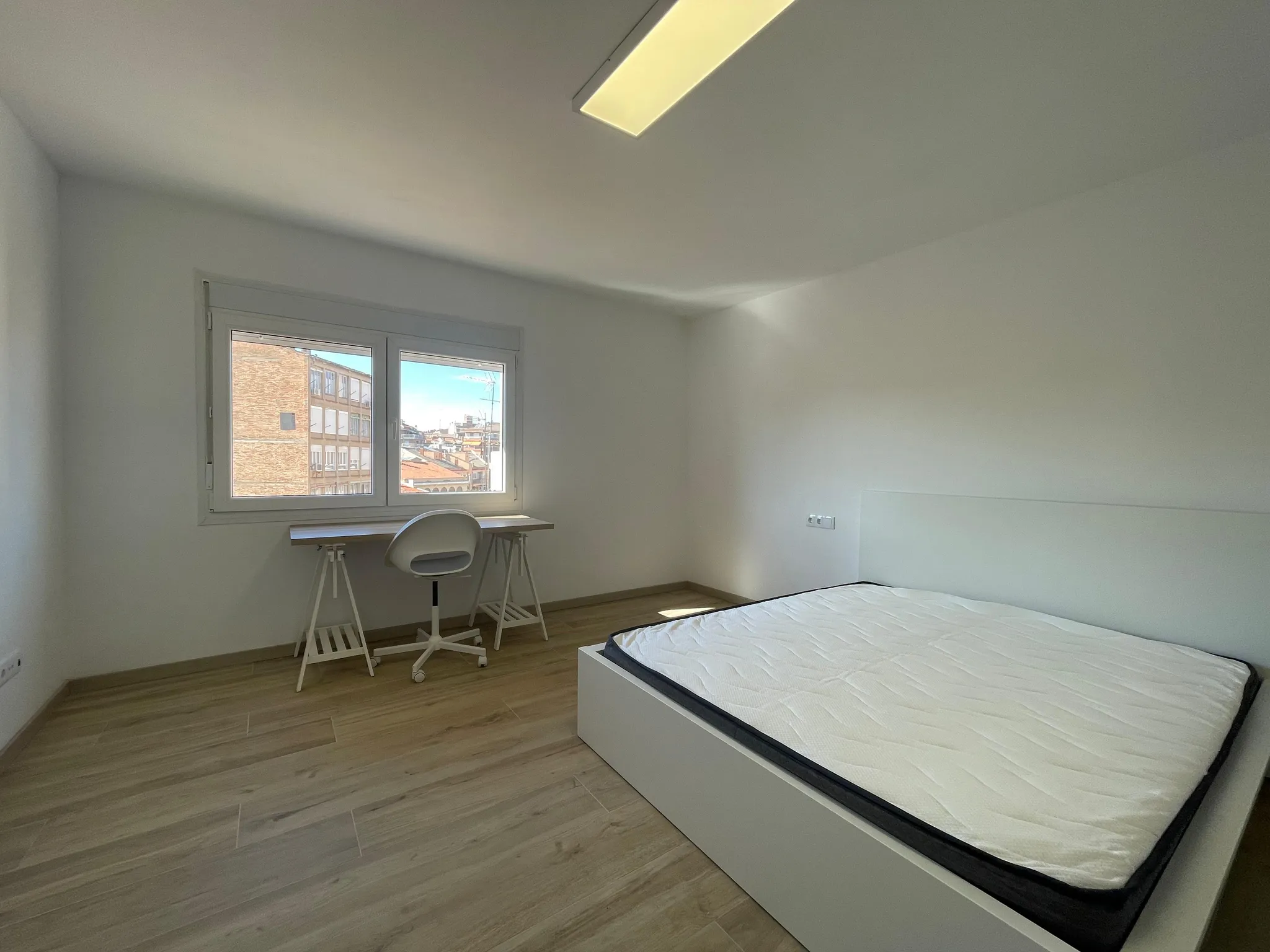 Bedroom with a mattress on a low bed frame, a small desk and chair under a window, white walls and wood-look floor.