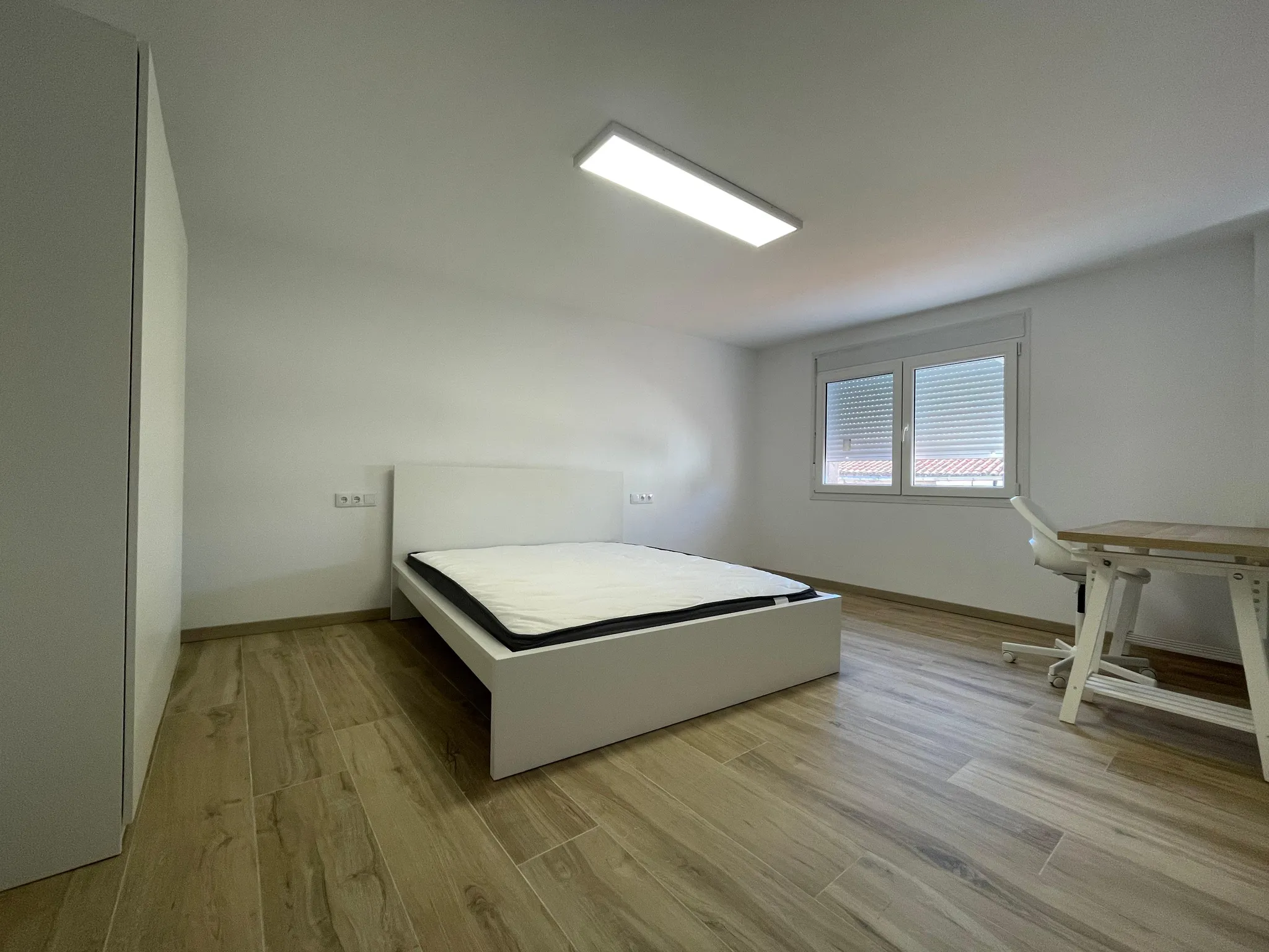 Bedroom with a single bed frame and mattress, window with closed blinds, small desk and chair, wardrobe at left, wood-look floor and ceiling light.