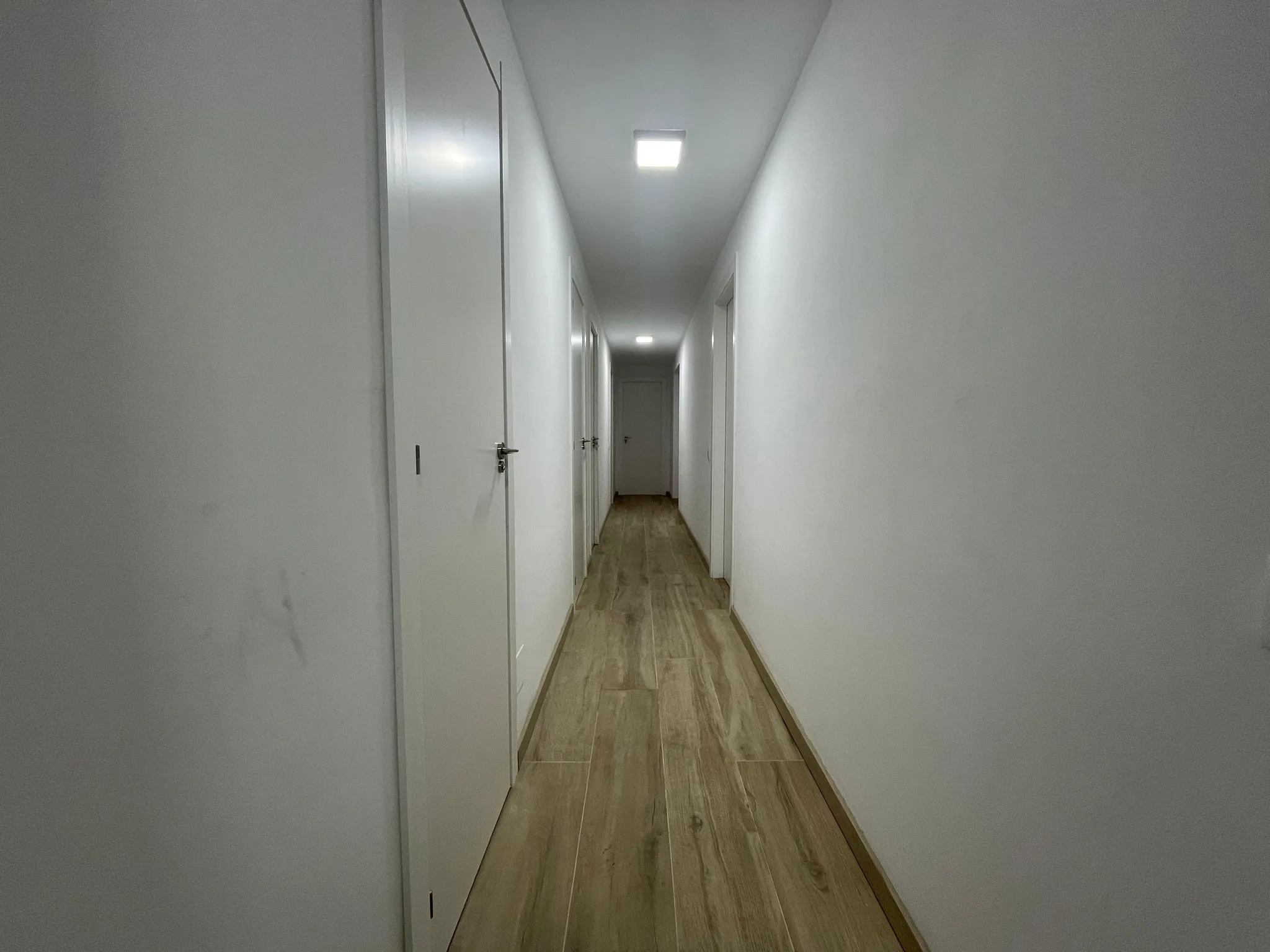Narrow interior hallway with white walls, multiple closed white doors on both sides and wood-look floor, ceiling lights visible.