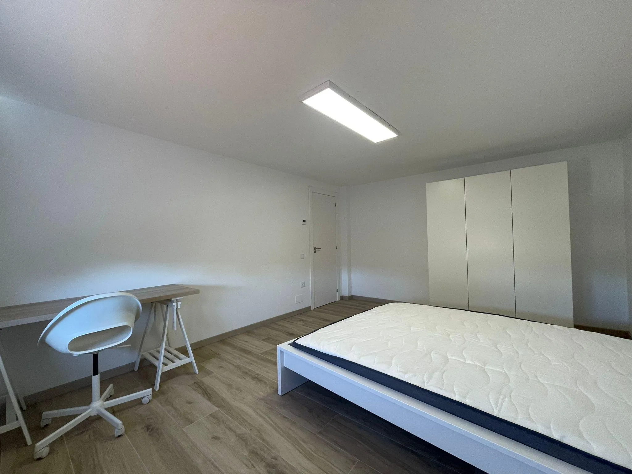 Bedroom with a bed frame and mattress, white wardrobe, desk with chair, wood-look floor and a ceiling light.