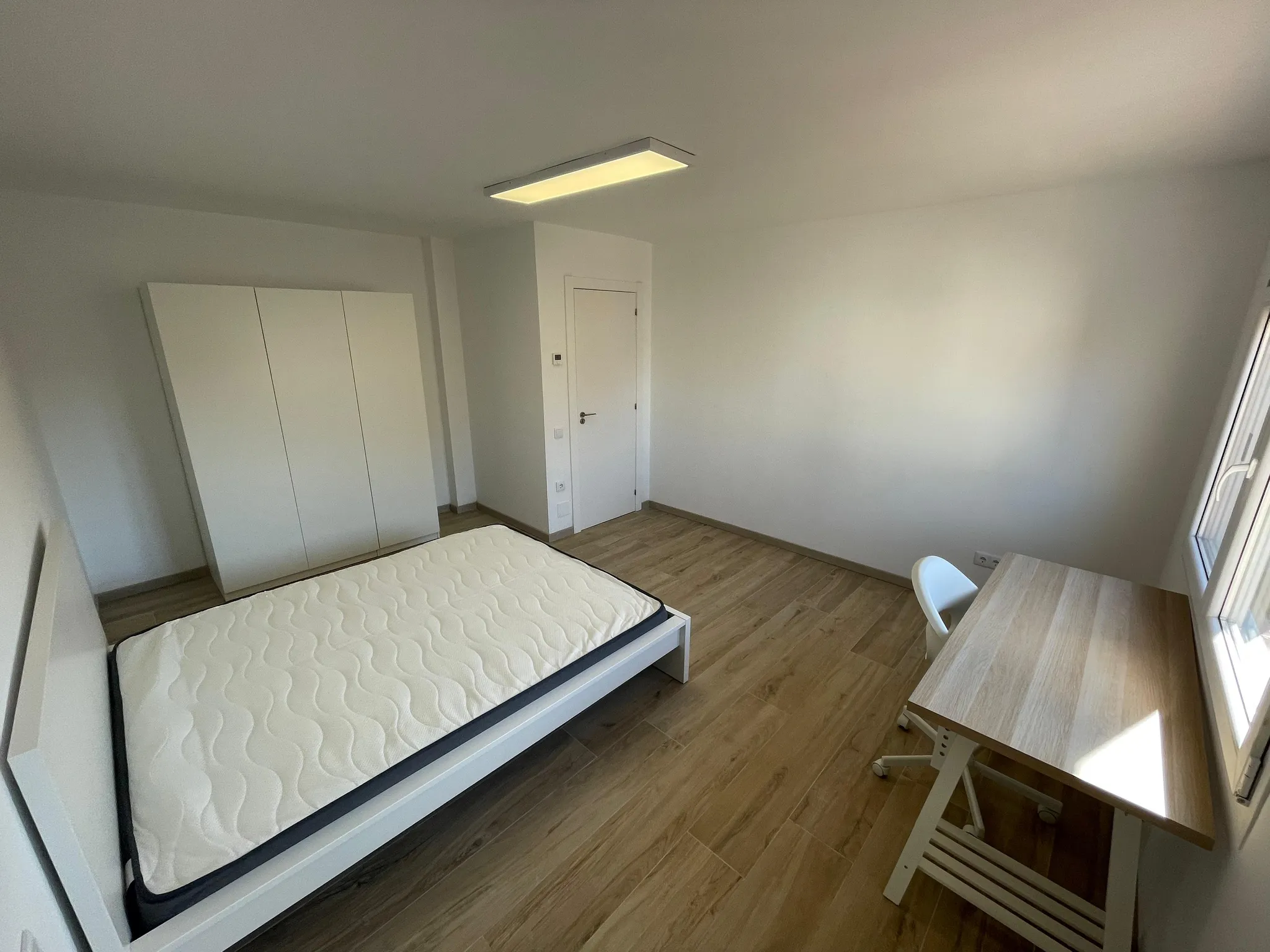 Bedroom with a single bed frame and mattress, wardrobe, desk and chair near a window, ceiling light and wood-look floor.