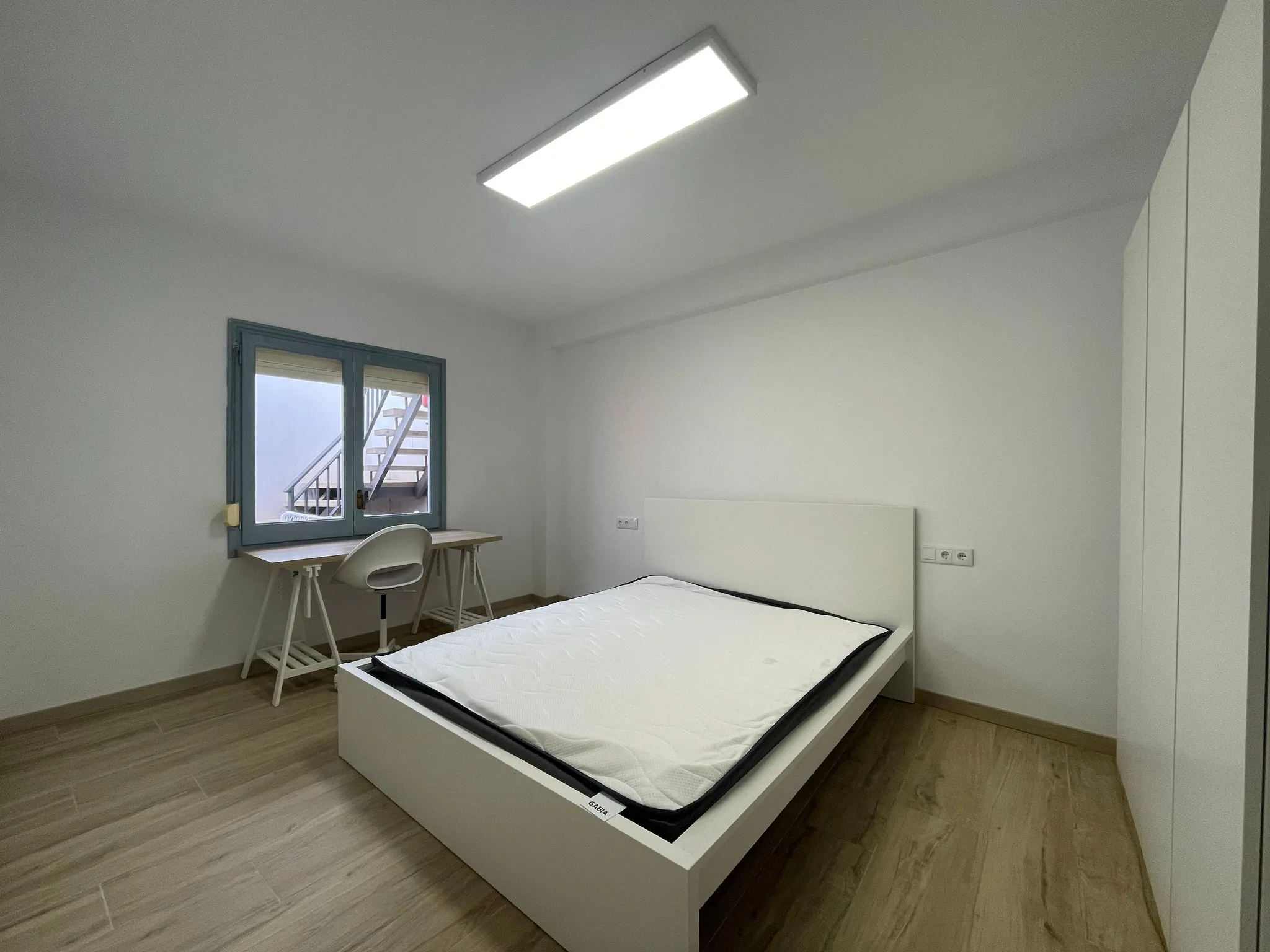 Bedroom with a single bed and mattress, a small desk and chair by a window, a wardrobe on the right and a ceiling light.