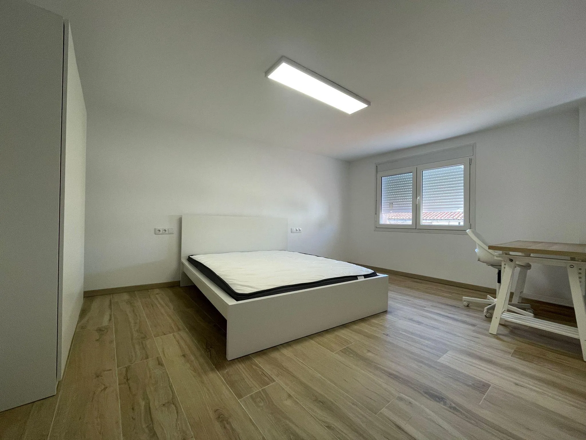 Bedroom with a single bed frame and mattress, a window with closed blinds, a desk and chair at right, wardrobe at left, ceiling light, and wood-look flooring.