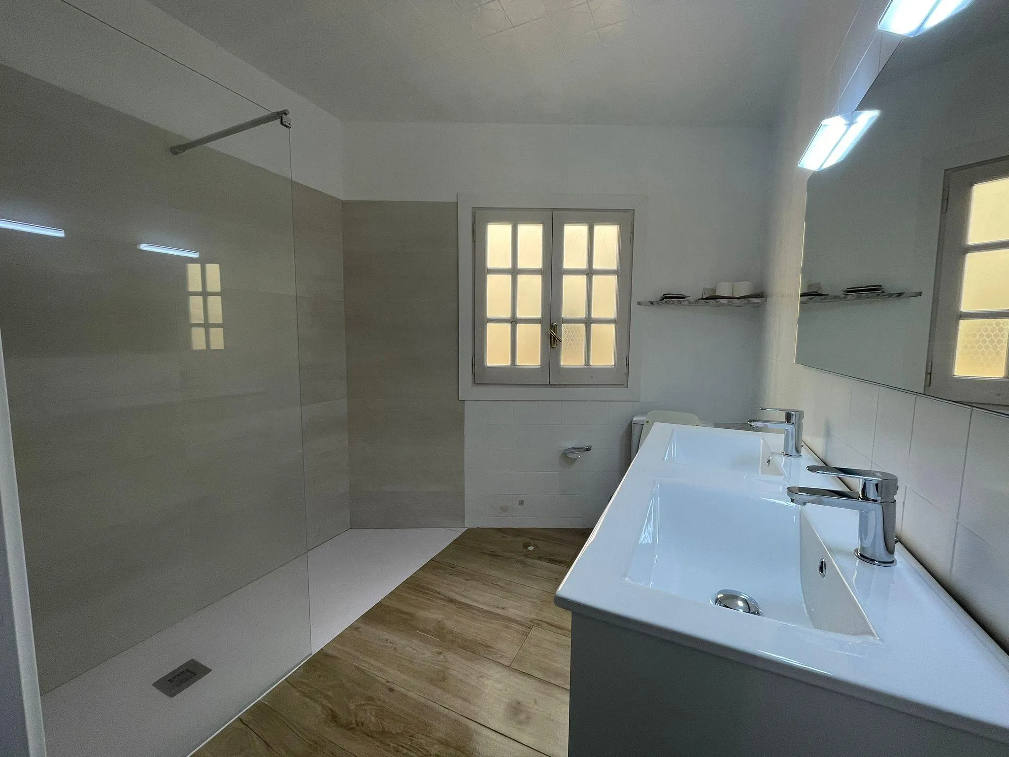 Bathroom with a double sink and faucets, large mirror, frosted window, and a walk-in shower area