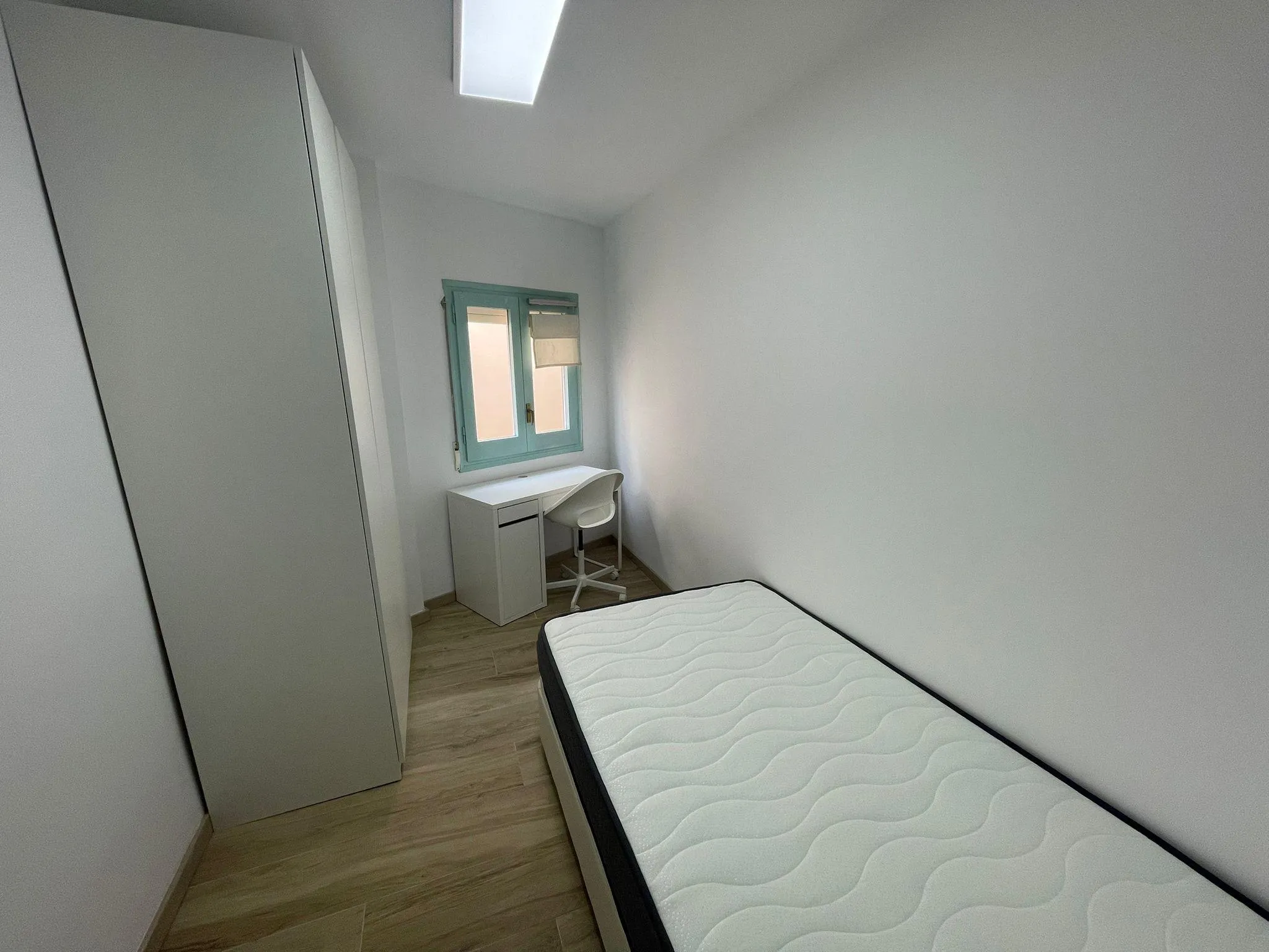 Small bedroom with a single bed, wardrobe, white desk and chair beside a window with a light-blue frame.