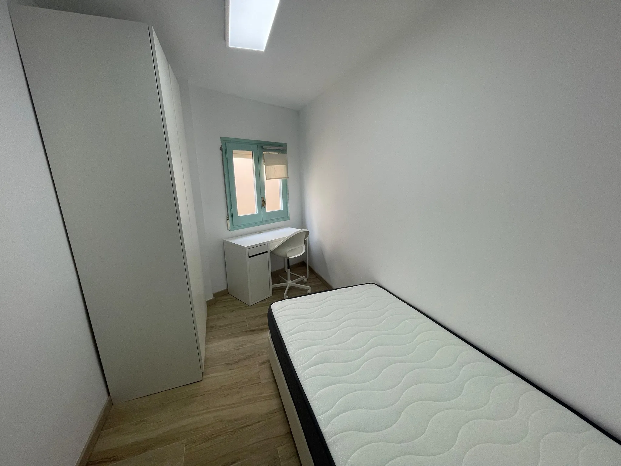 Small bedroom with a single mattress on a bed frame, wardrobe on the left, a desk and chair under a small window with a blind, and a rectangular skylight in the ceiling.