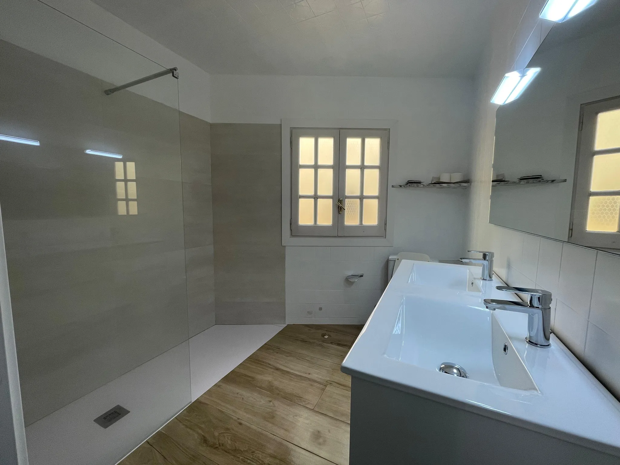 Bathroom with a walk-in glass shower, double sink vanity with chrome faucets, wall mirror, small shelf and frosted window.