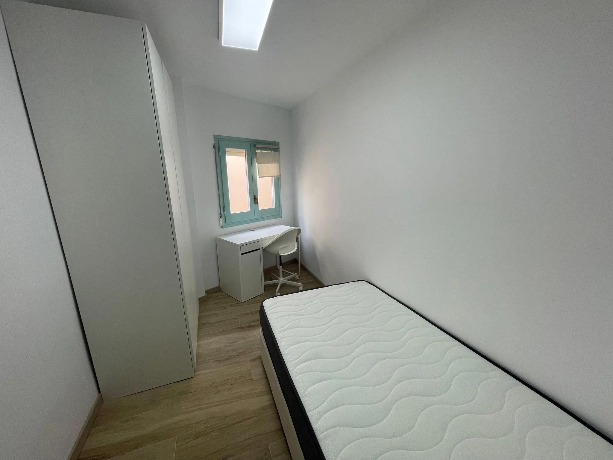 Small bedroom with a single mattress on a bed frame, wardrobe on the left, and a small desk with a chair under a window.